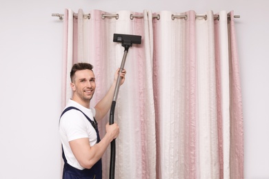 Photo of Male worker removing dust from curtains with professional vacuum cleaner indoors