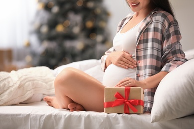 Happy pregnant woman with Christmas gift box on bed at home, closeup. Expecting baby