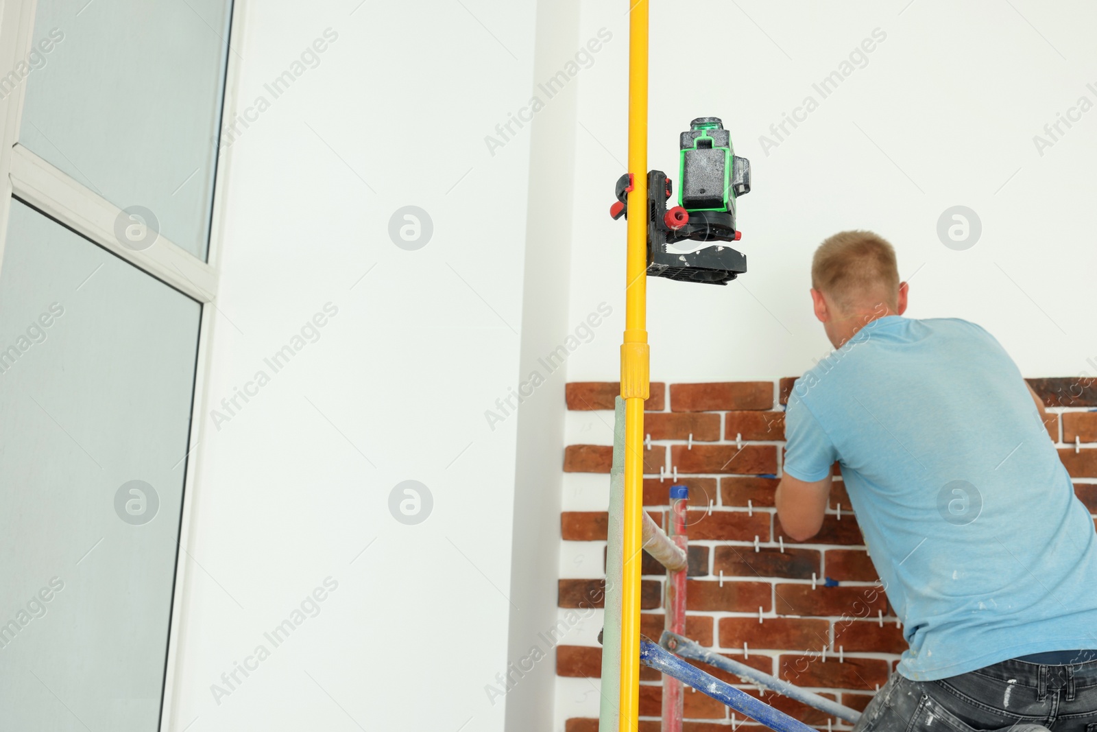 Photo of Professional builder using cross line laser level while gluing decorative bricks on wall, back view. Tiles installation process