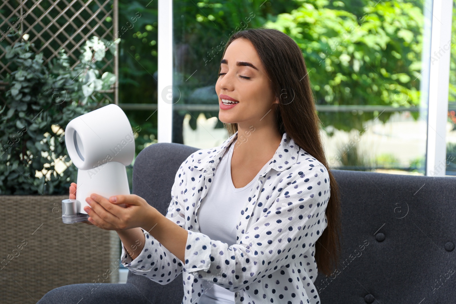 Photo of Woman enjoying air flow from portable fan at home. Summer heat