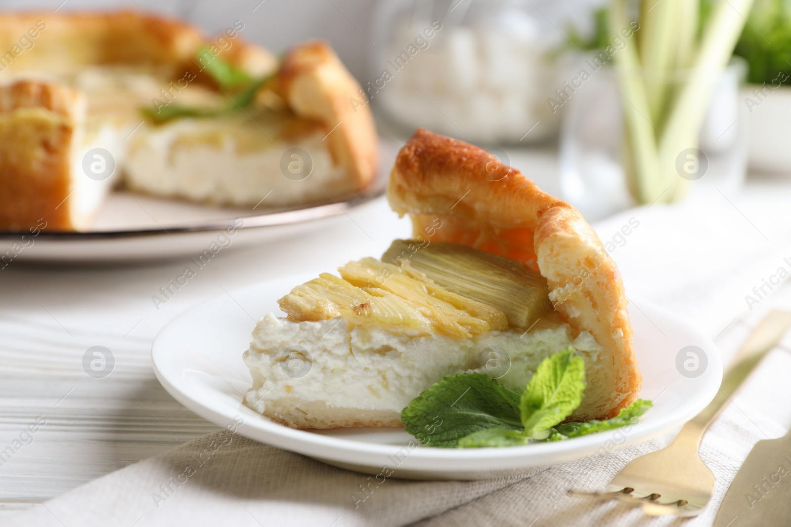 Photo of Piece of freshly baked rhubarb pie with cream cheese, mint leaves and cutlery on white wooden table, closeup