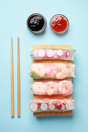 Photo of Different delicious spring rolls, chopsticks and sauces on light blue background, flat lay