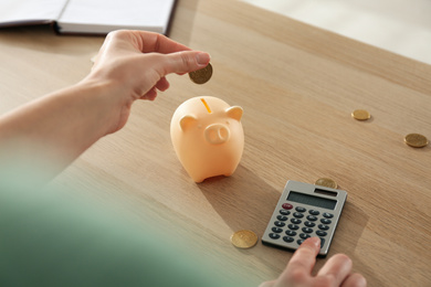 Photo of Woman putting money into piggy bank and using calculator at wooden table indoors, closeup