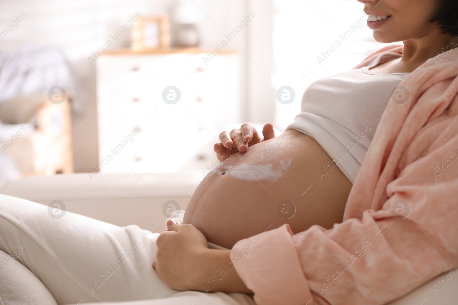 Photo of Young pregnant woman applying cosmetic product on belly at home, closeup