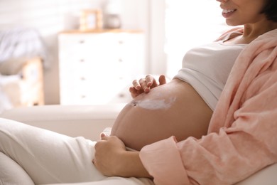 Young pregnant woman applying cosmetic product on belly at home, closeup