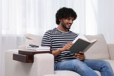 Photo of Happy man reading book on sofa with wooden armrest table at home