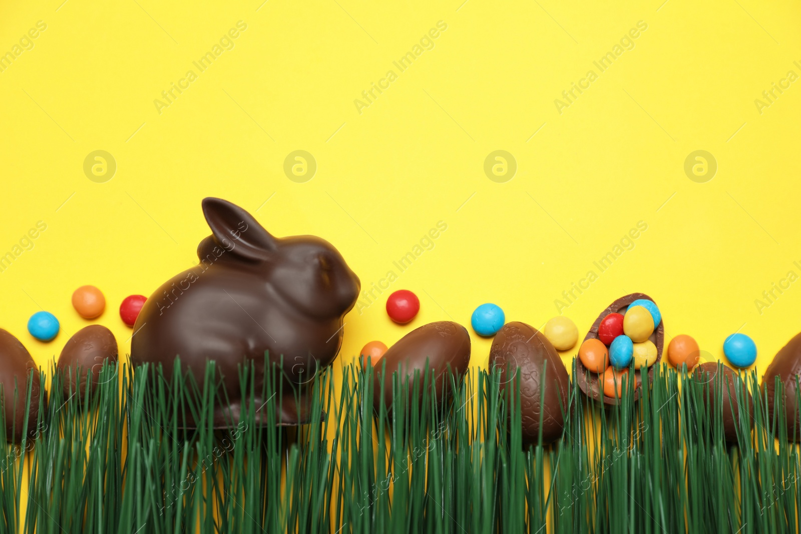 Photo of Chocolate Easter bunny, eggs and candies with green grass on yellow background. Space for text