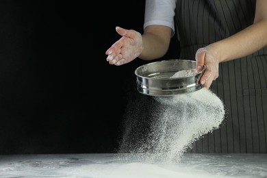 Photo of Woman sieving flour at table against black background, closeup. Space for text