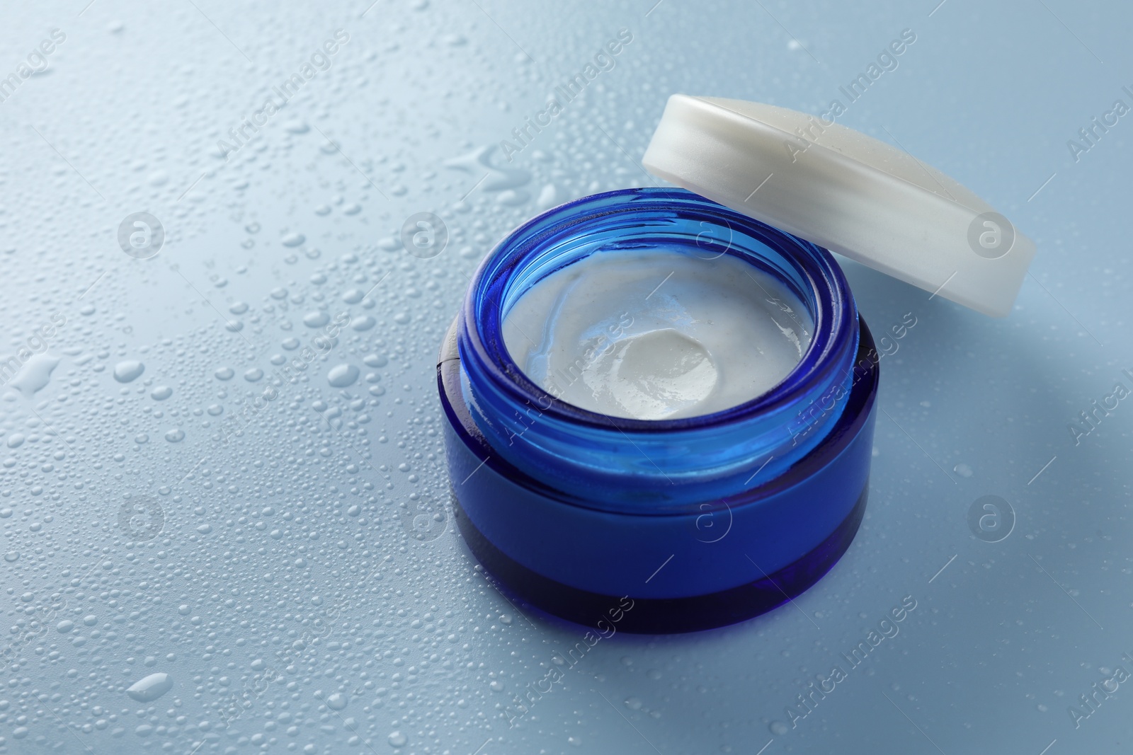 Photo of Moisturizing cream in open jar on light blue background with water drops, closeup. Space for text