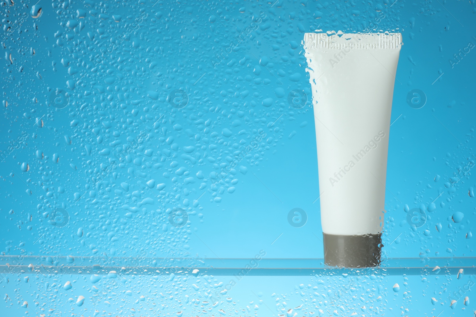 Photo of Tube with moisturizing cream on light blue background, view through wet glass. Space for text