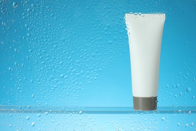 Tube with moisturizing cream on light blue background, view through wet glass. Space for text