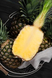 Photo of Whole and cut ripe pineapples in metal basket on black wooden table, top view