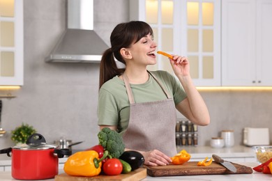 Young housewife tasting cut bell pepper while cooking at white table in kitchen