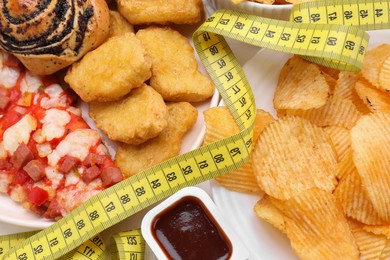 Different unhealthy food and measuring tape on white table, top view. Weight loss concept