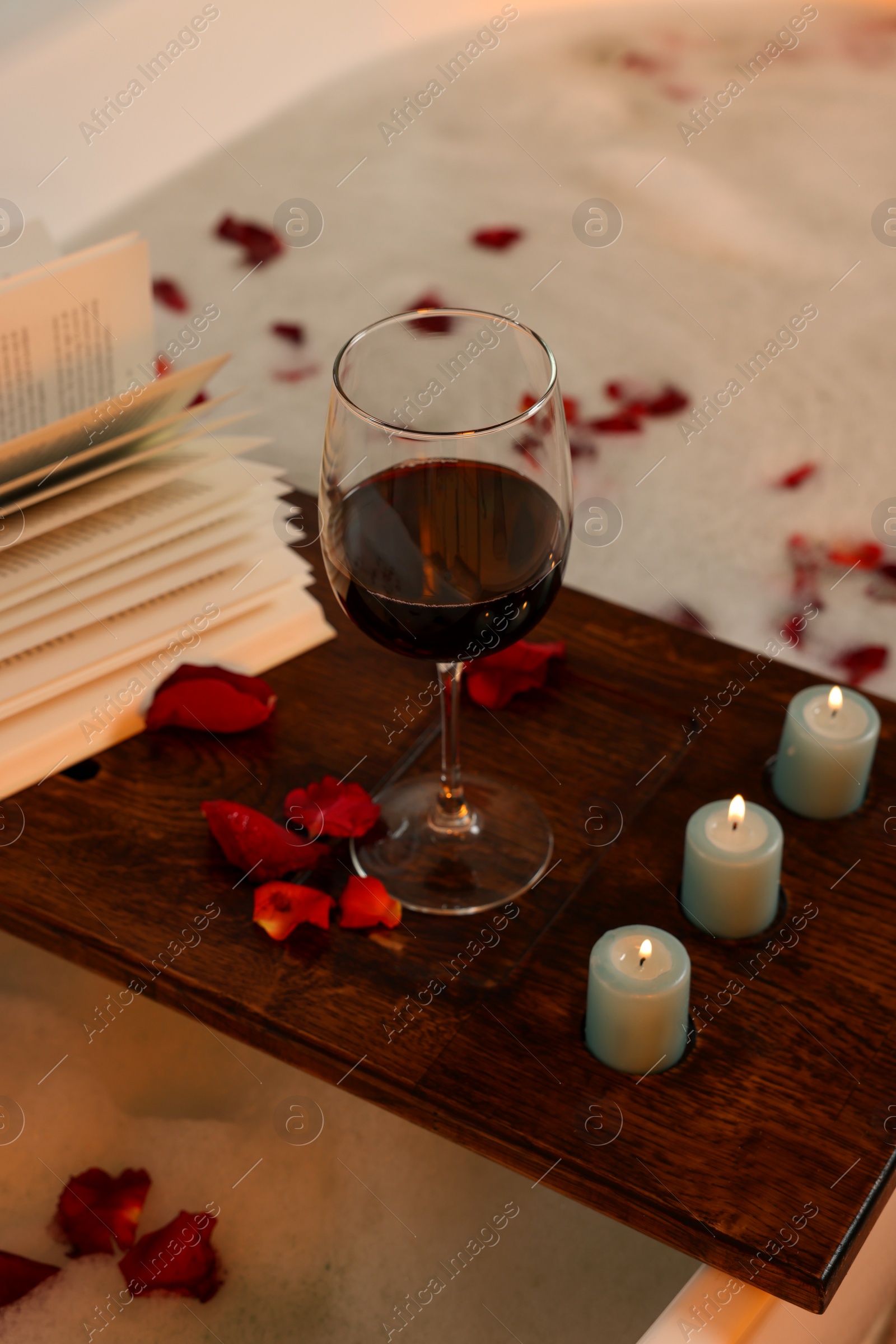 Photo of Wooden board with glass of wine, book, burning candles and rose petals on bath tub