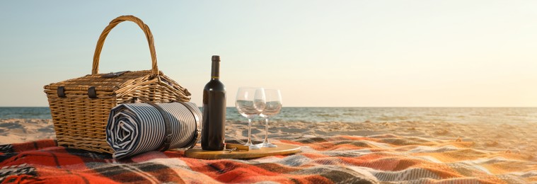 Image of Blanket with picnic basket, bottle of wine and glasses on sandy beach near sea, space for text. Banner design