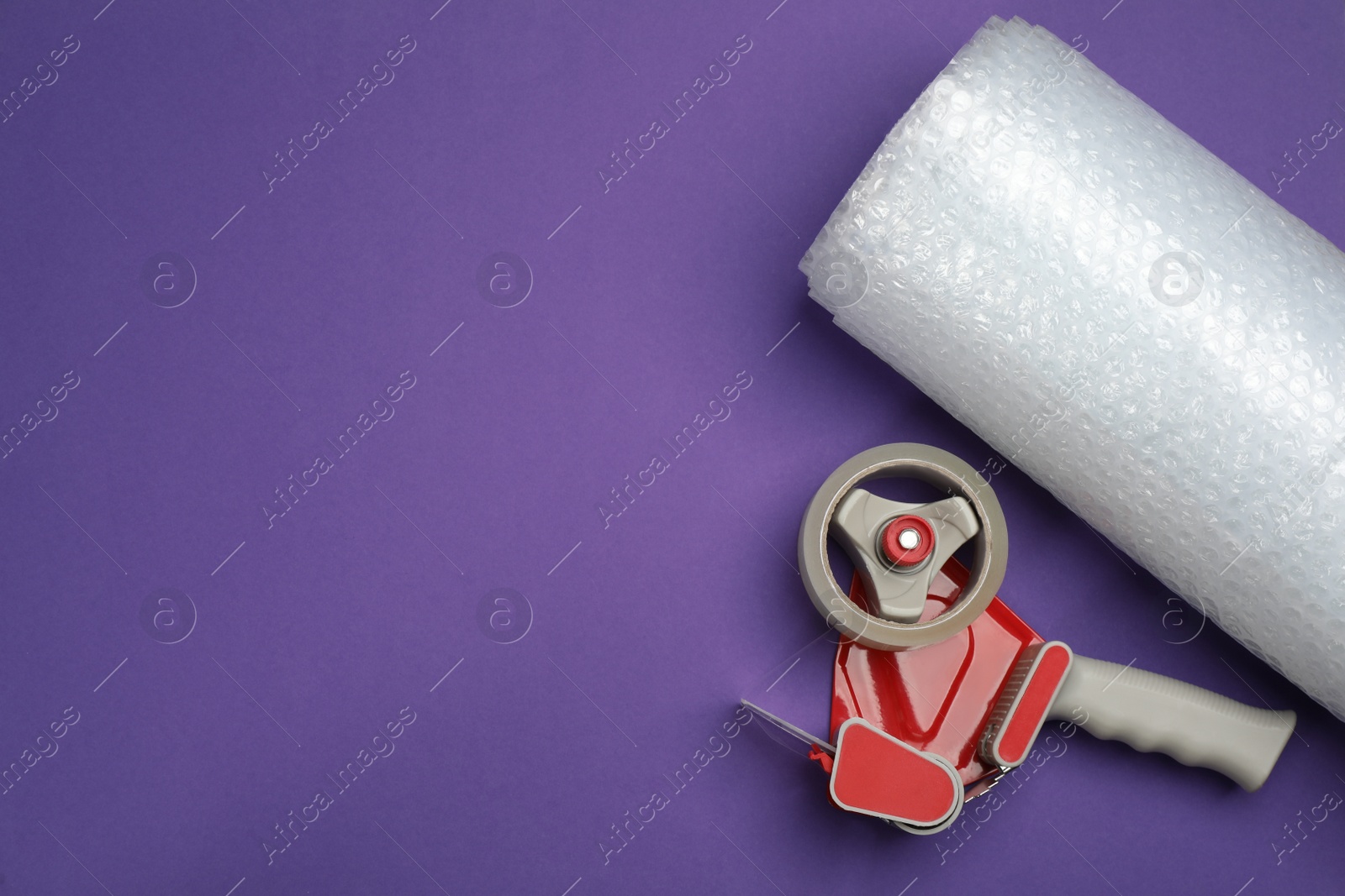 Photo of Bubble wrap roll and tape dispenser on purple background, flat lay. Space for text