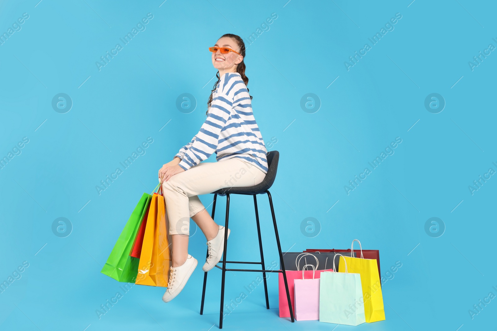 Photo of Happy woman in stylish sunglasses holding colorful shopping bags on stool against light blue background