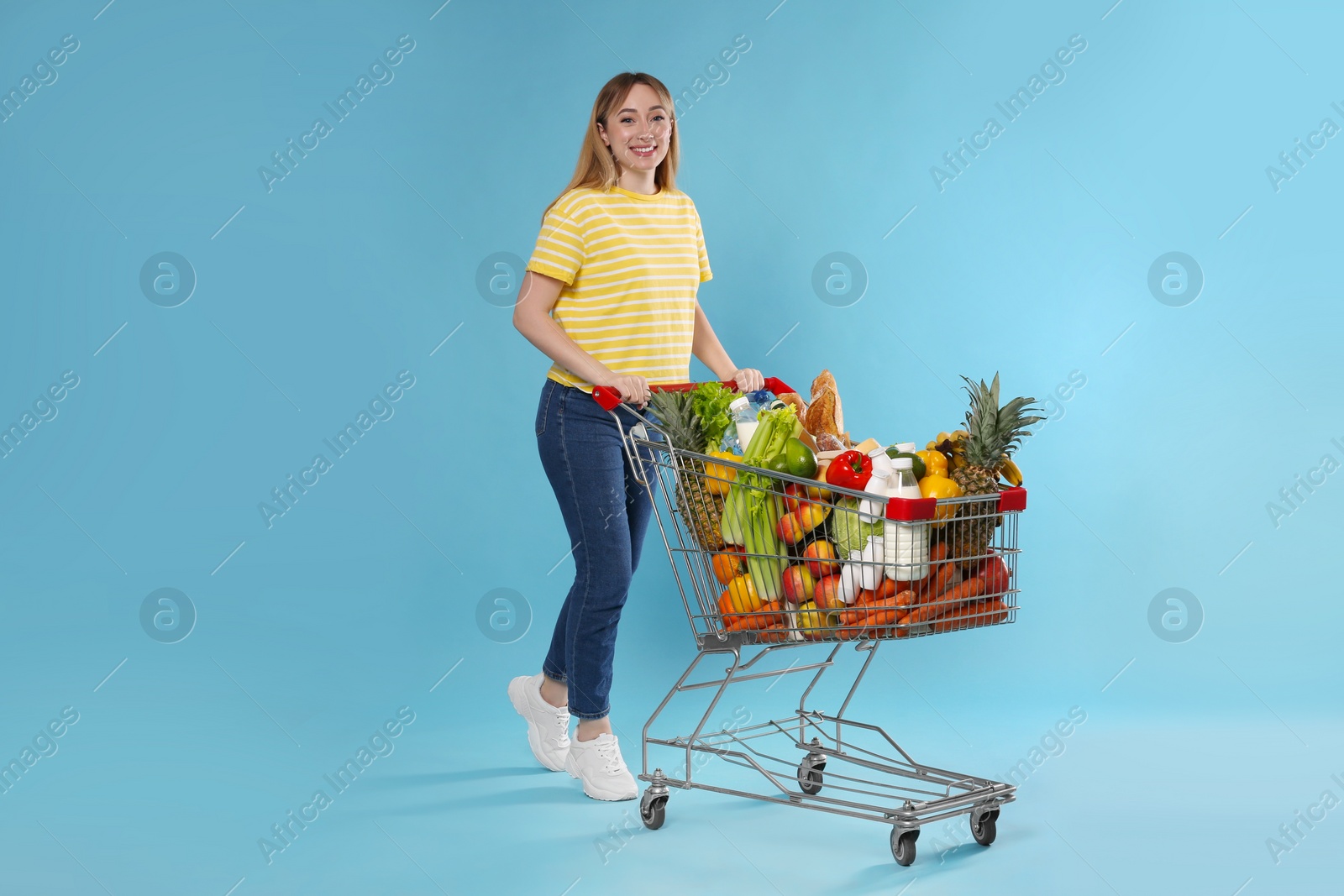 Photo of Young woman with shopping cart full of groceries on light blue background