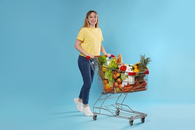Photo of Young woman with shopping cart full of groceries on light blue background