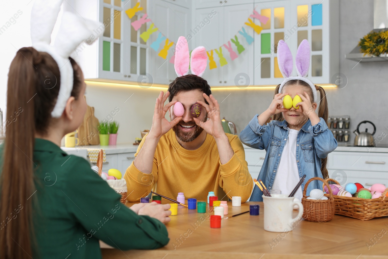 Photo of Father and daughter covering eyes with Easter eggs at table in kitchen. Family having fun