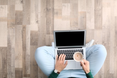 Young man with laptop and cup of coffee sitting on floor, top view