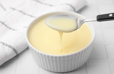 Photo of Condensed milk flowing down from spoon into bowl on white tiled table, closeup