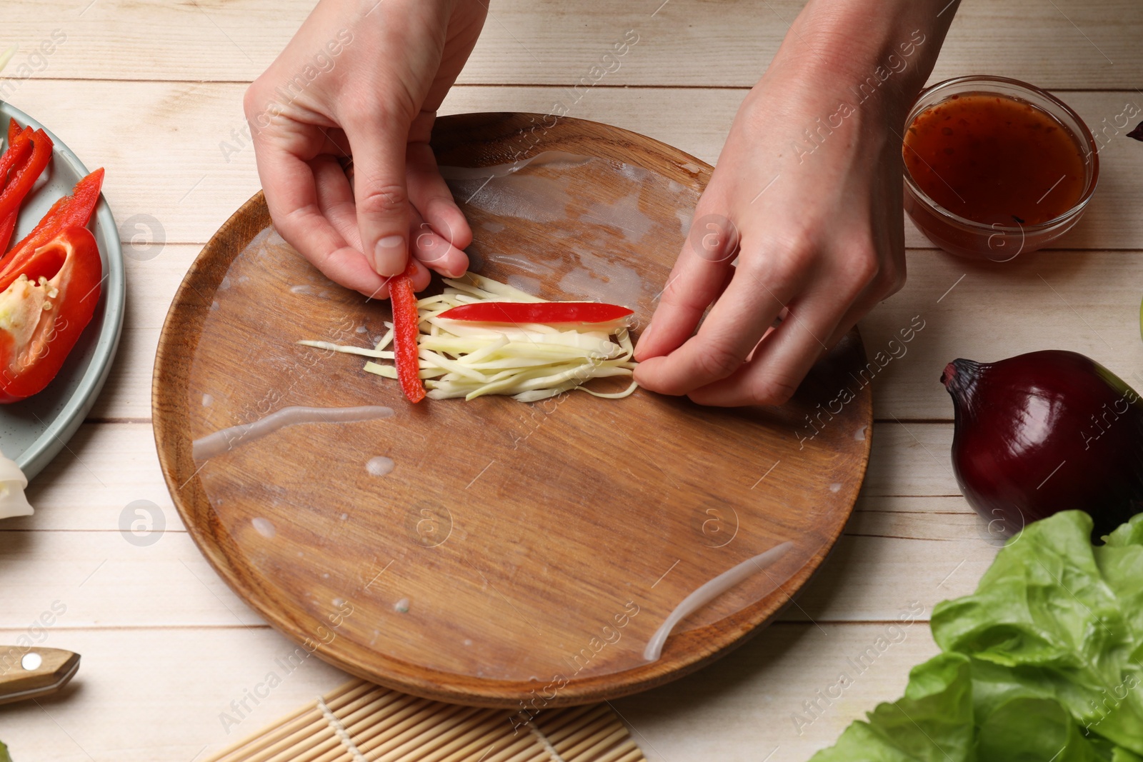 Photo of Making delicious spring rolls. Woman wrapping fresh vegetables into rice paper at wooden table, closeup