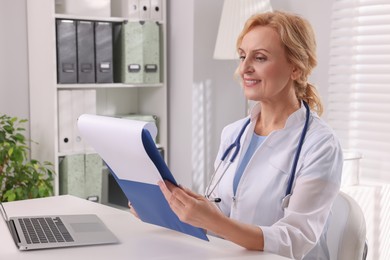 Photo of Doctor with laptop and clipboard at white table in clinic. Patient consultation