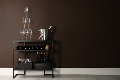 Rack with bottles of wine and glasses near brown wall, space for text
