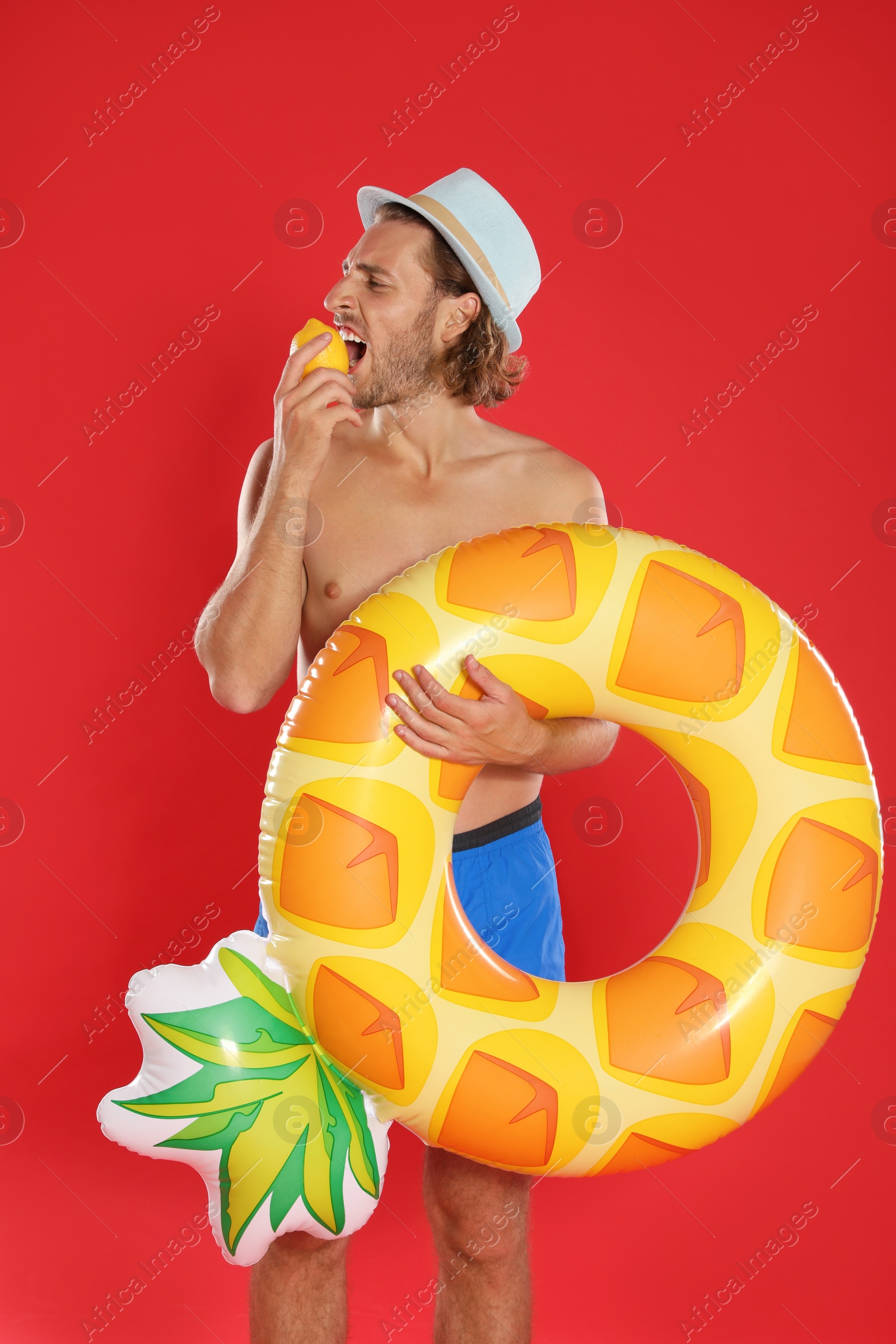 Photo of Attractive young man in swimwear with pineapple inflatable ring eating lemon on red background