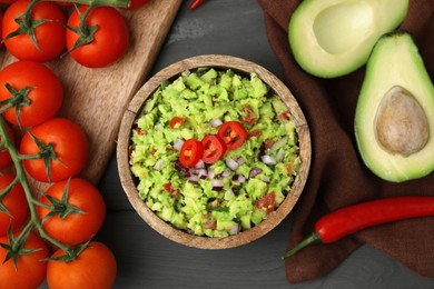 Bowl of delicious guacamole and ingredients on grey wooden table, flat lay