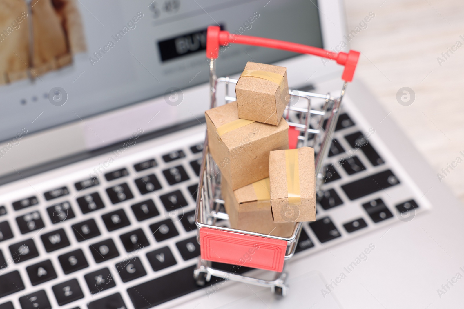 Photo of Internet store. Small cardboard boxes, shopping cart and laptop on table, closeup. Space for text