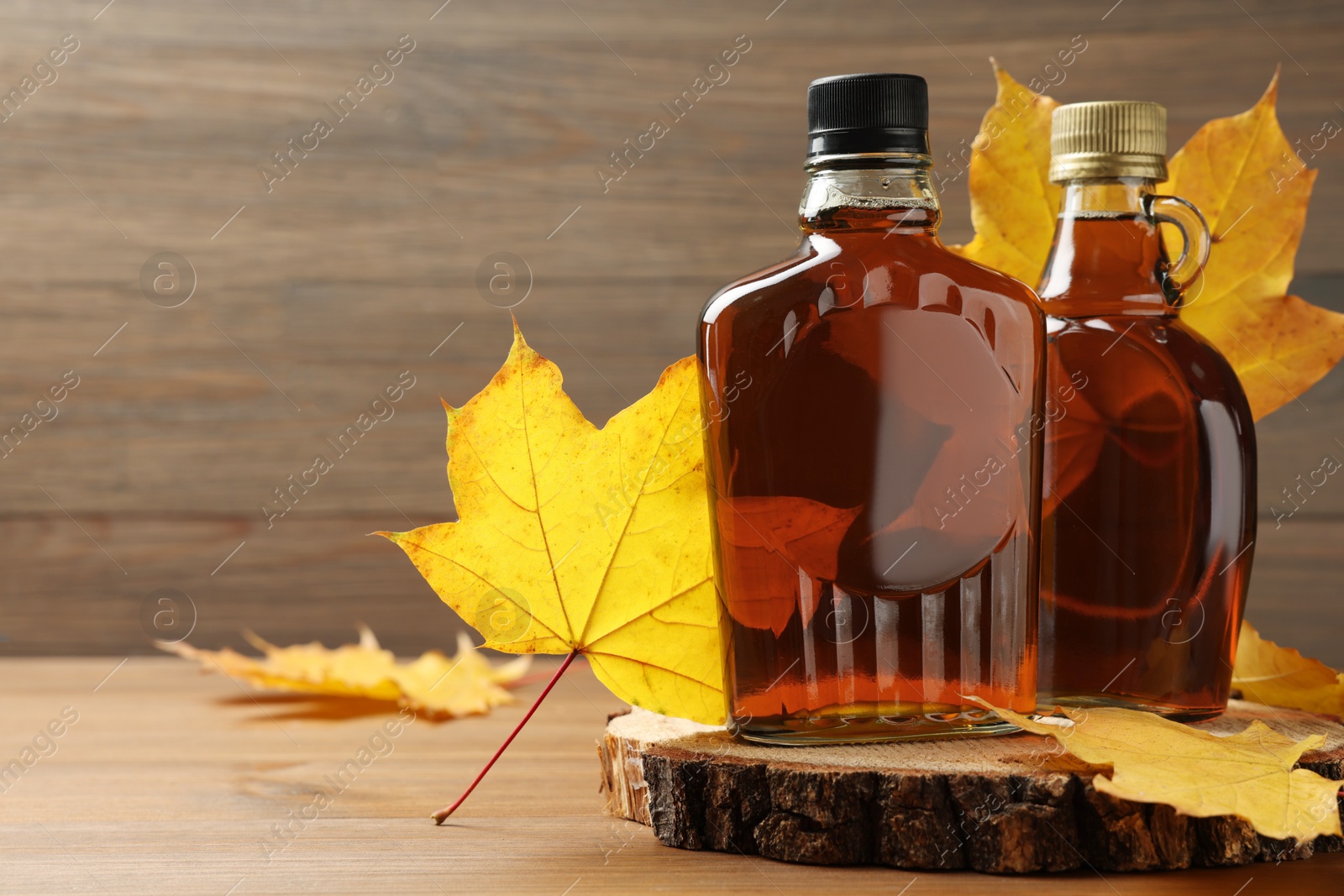 Photo of Bottles of tasty maple syrup and dry leaves on wooden table, space for text