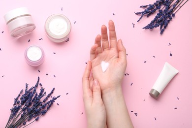 Woman applying hand cream and lavender flowers on pink background, top view