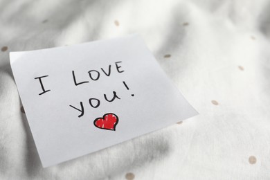 Sticky note with phrase I love you! on bed, closeup