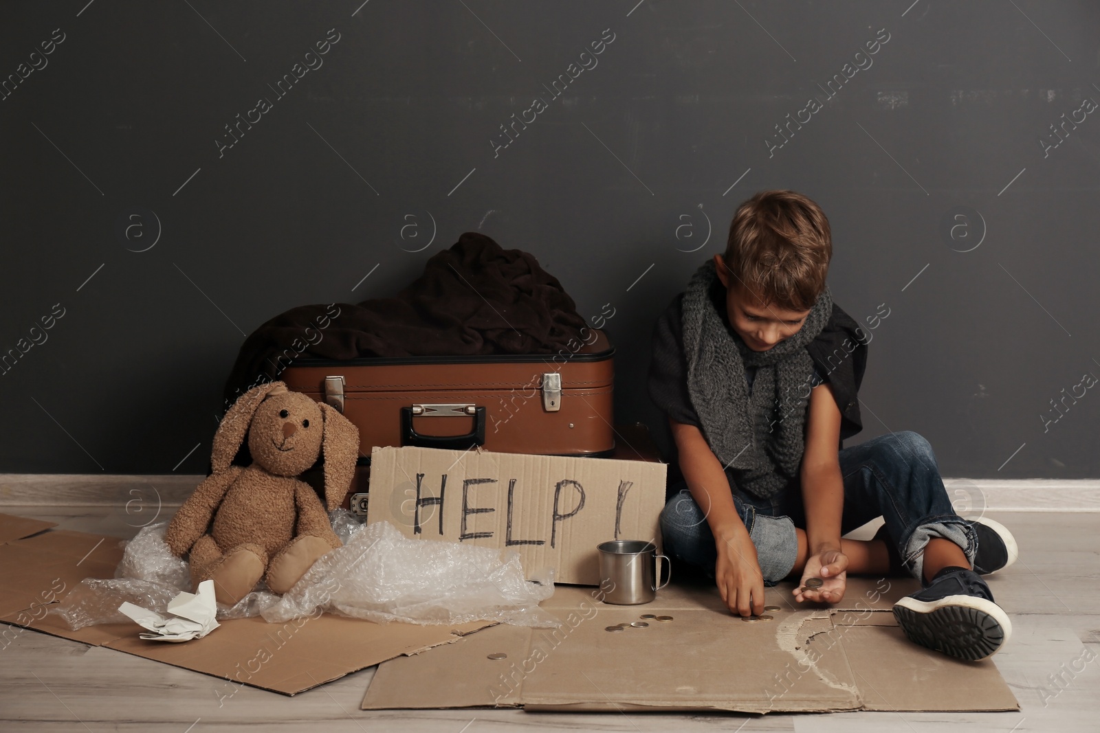 Photo of Poor boy with suitcases and toy counting coins on floor near dark wall