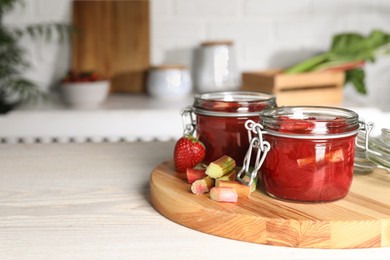 Jars of tasty rhubarb jam, cut stems and strawberry on white wooden table. Space for text