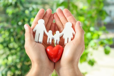 Photo of Young man holding wooden family figure and red heart in his hands against blurred background, closeup