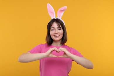 Photo of Happy woman with bunny ears showing heart gesture with hands on orange background