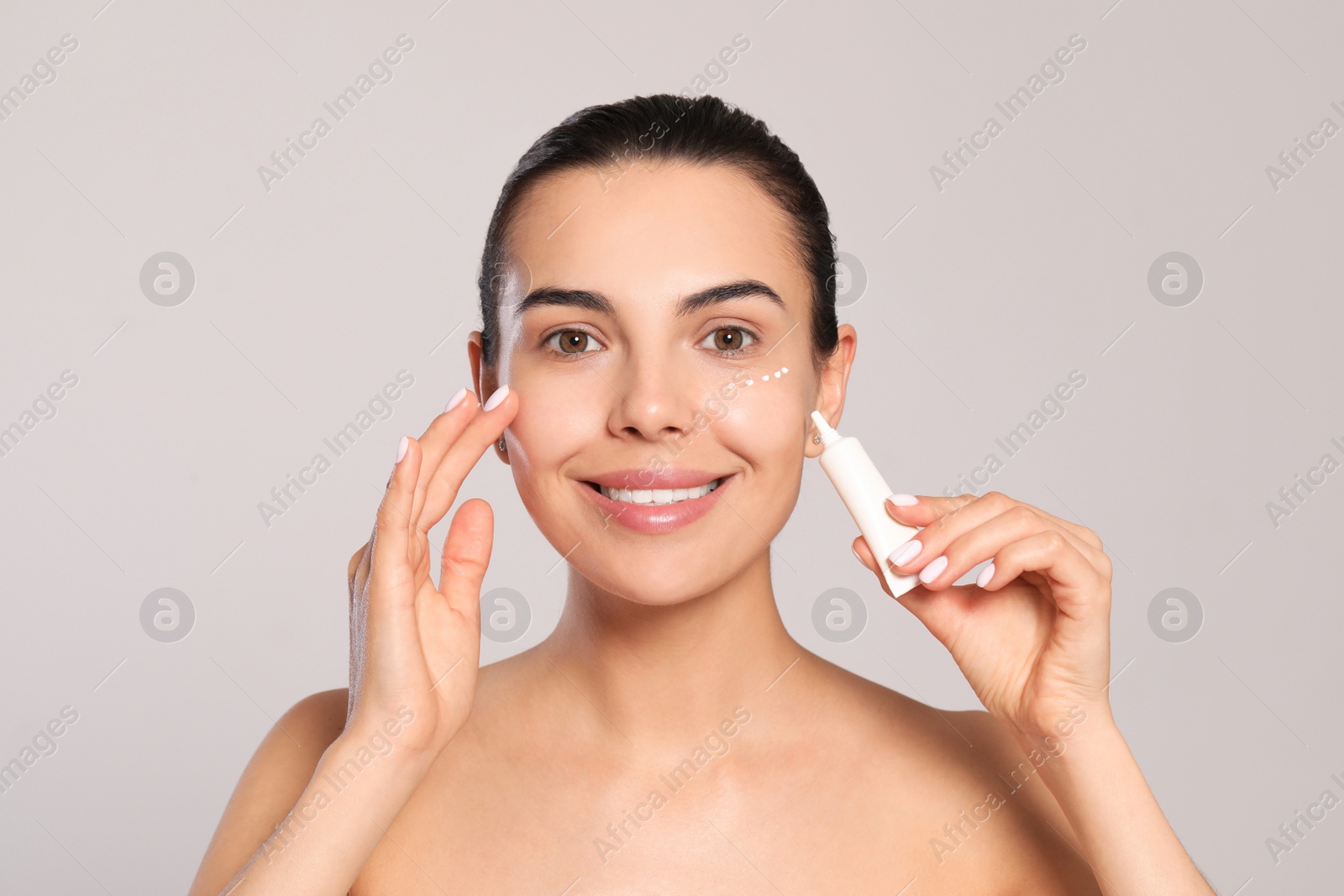 Photo of Beautiful young woman with cream on skin under eye against light grey background
