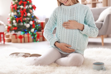 Photo of Happy pregnant woman sitting on floor in room decorated for Christmas