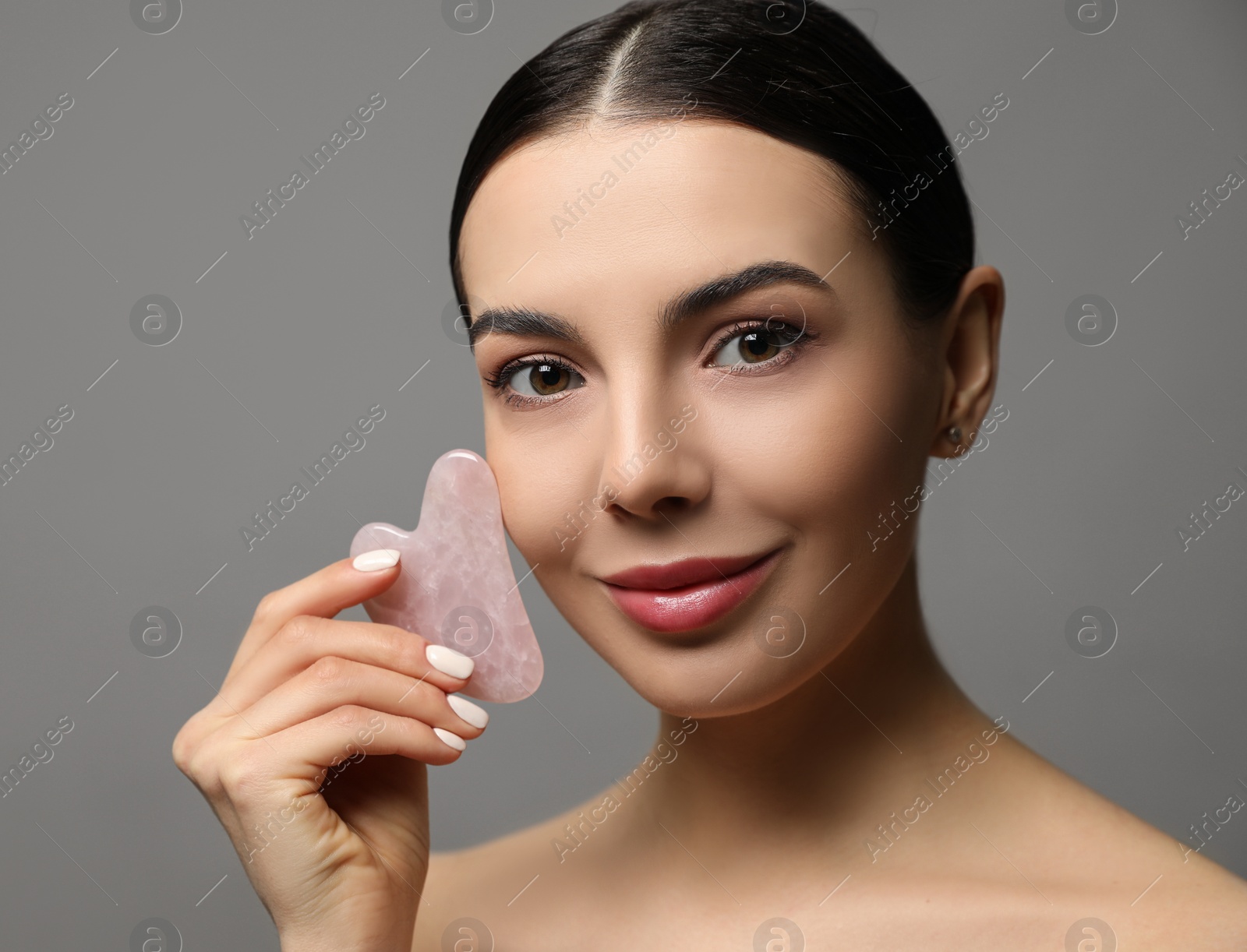 Photo of Beautiful young woman doing facial massage with gua sha tool on grey background, closeup