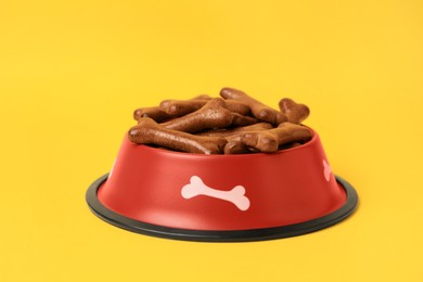 Red bowl with bone shaped dog cookies on yellow background, closeup
