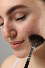 Photo of Beautiful woman with freckles applying makeup with brush on grey background, closeup