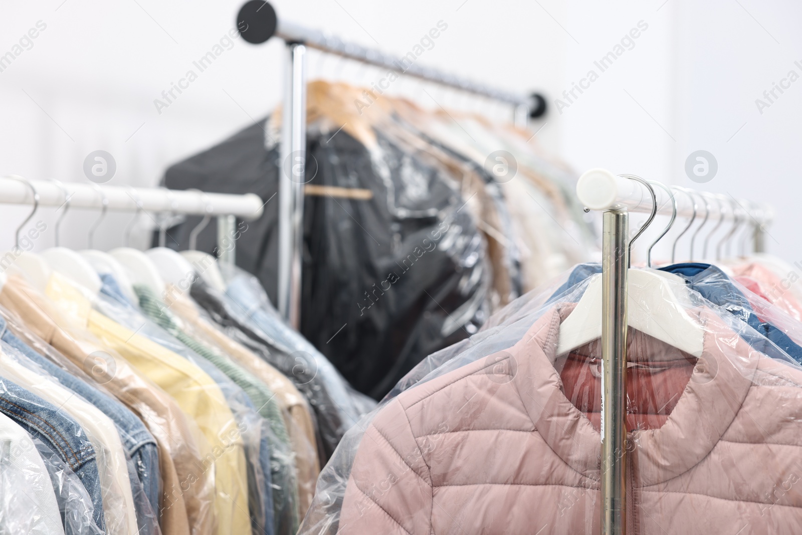 Photo of Dry-cleaning service. Many different clothes in plastic bags hanging on rack indoors, closeup