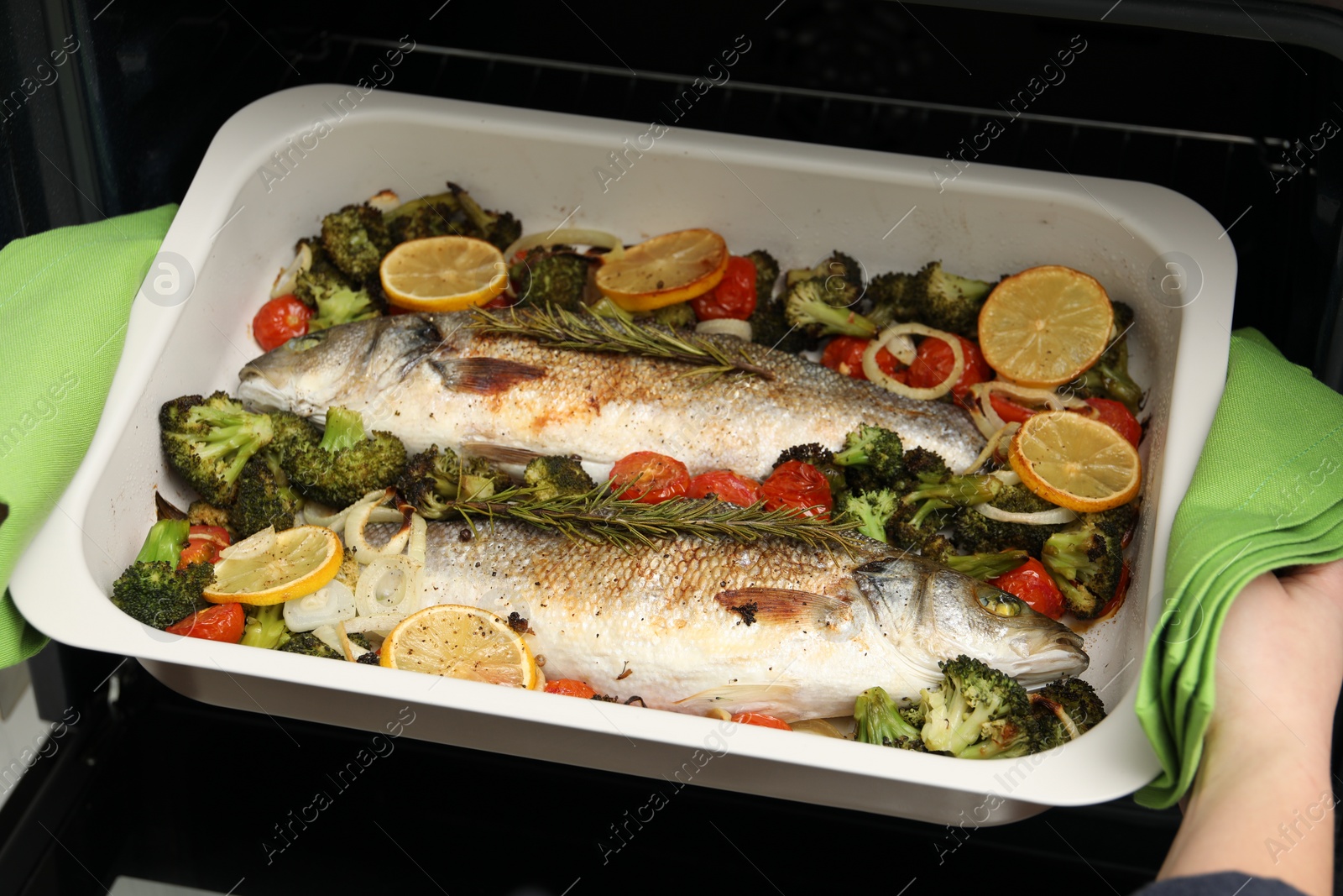 Photo of Woman taking baking dish with delicious fish and vegetables from oven, closeup