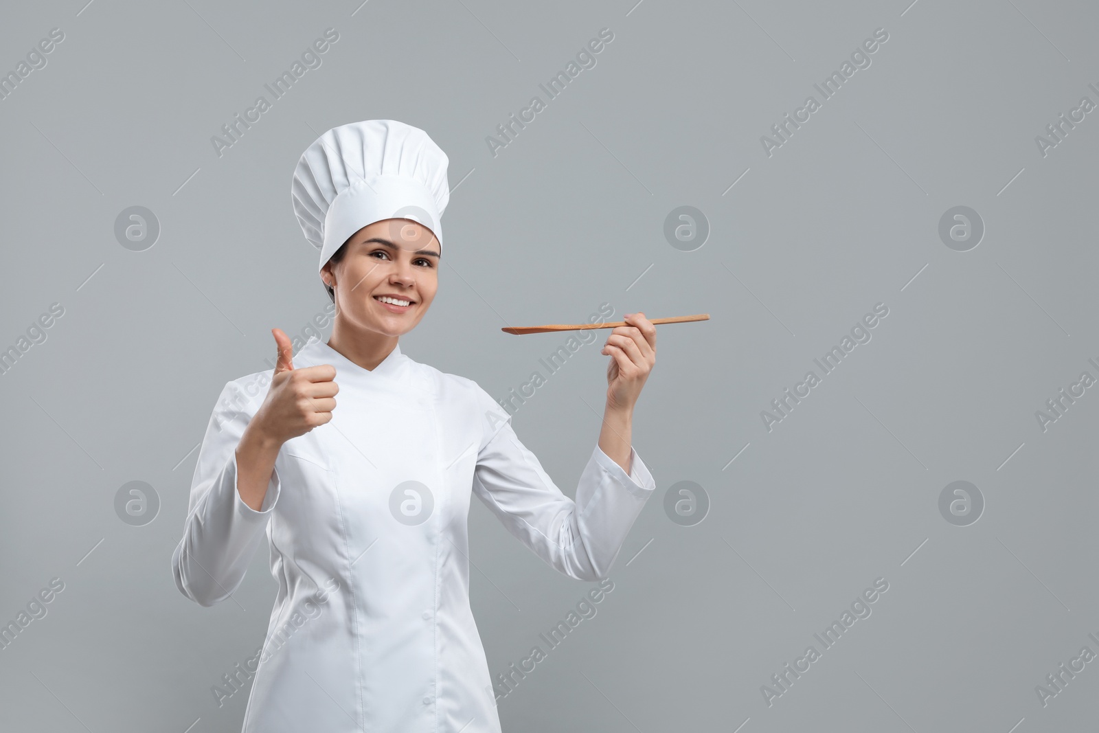 Photo of Happy female chef with wooden spoon showing thumbs up on light grey background. Space for text