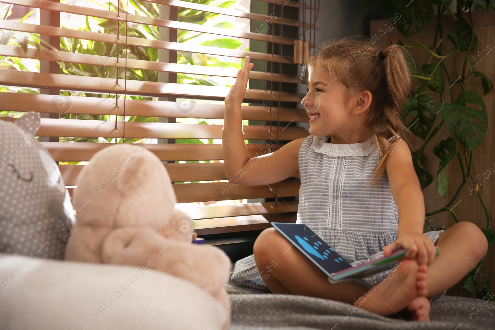 Photo of Cute little girl reading book near window at home