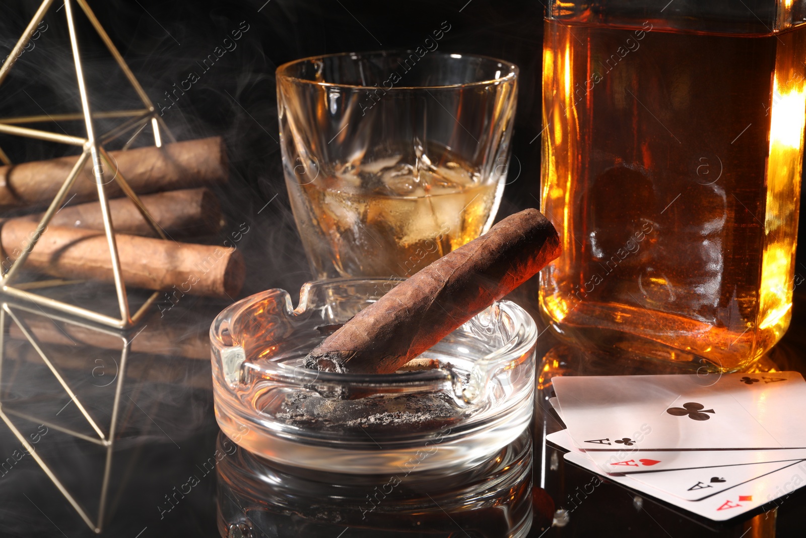 Photo of Smoldering cigar, ashtray, playing cards and whiskey on black mirror surface